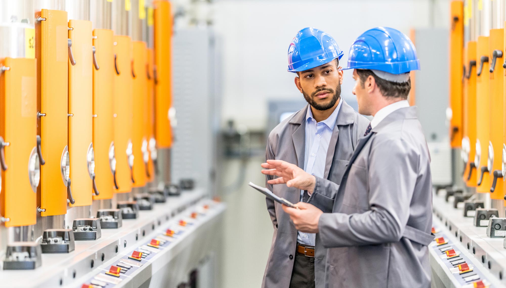 Deux professionnels en environnement industriel examinent une tablette devant des armoires électriques et équipements techniques. La scène illustre un échange autour de la performance, de la sécurité ou de la conformité des installations.